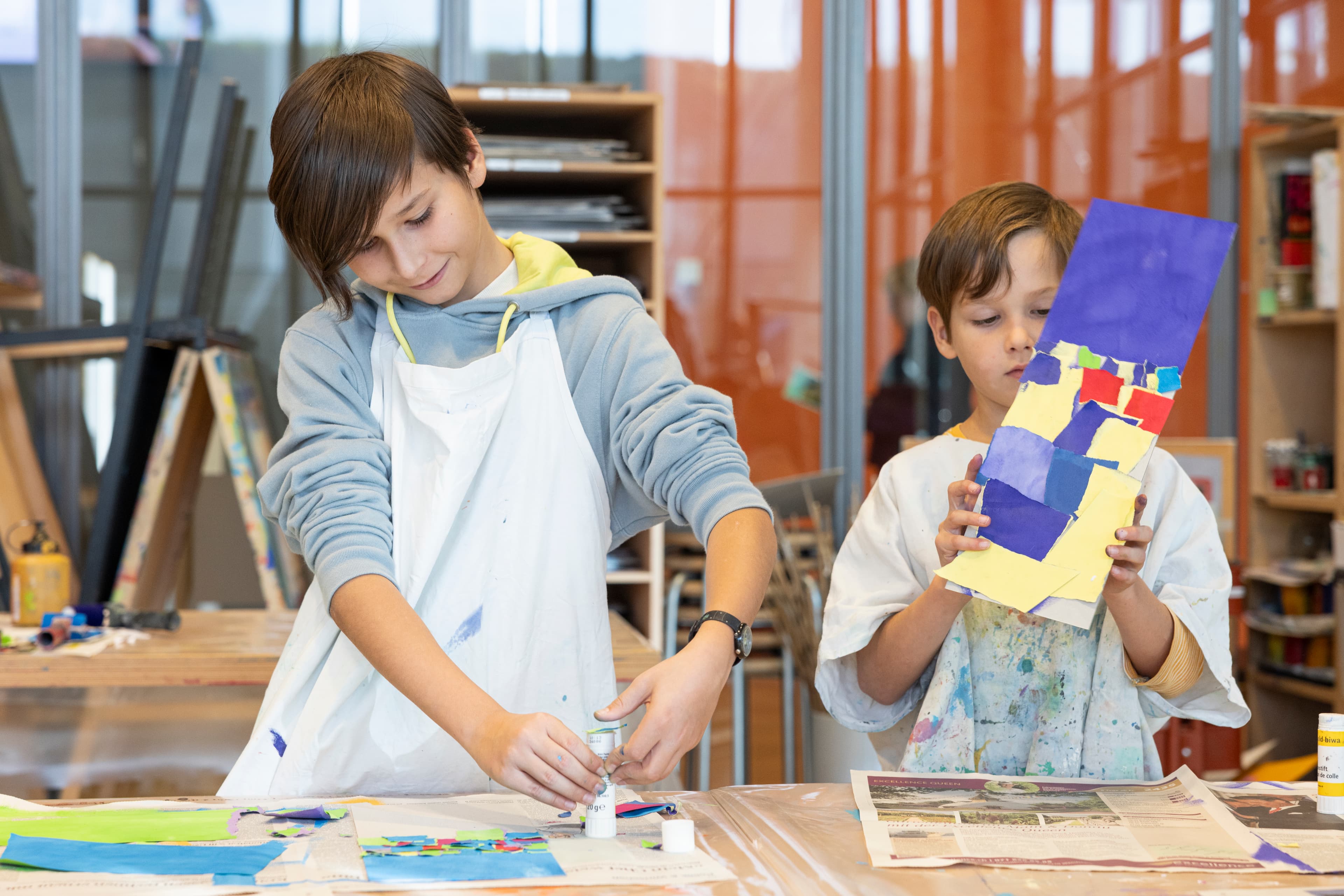 Zwei Kinder im Alter von 6 und 9 Jahren tragen Malschürzen und sind frontal zu sehen, wie sie kleine Kunstwerke im Atelier gestalten.