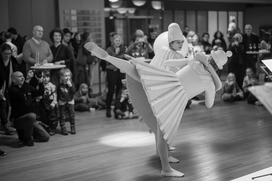Zwei Tänzerinnen in Kostümen, die dem Triadischen Ballett von Oskar Schlemmer nachempfunden sind, tanzen inmitten zahlreicher Besucher:innen.