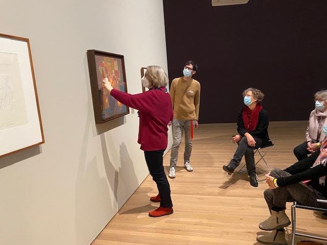 Eine Frau steht nahe einem Bild von Paul Klee und deutet auf ein Element des Werks. Im Hintergrund sind mehrere Menschen zu sehen, die aktiv zuhören.